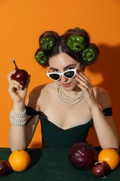 Photo of Young woman with peppers in her hair, sunglasses, fruits and vegetables at green table on orange background