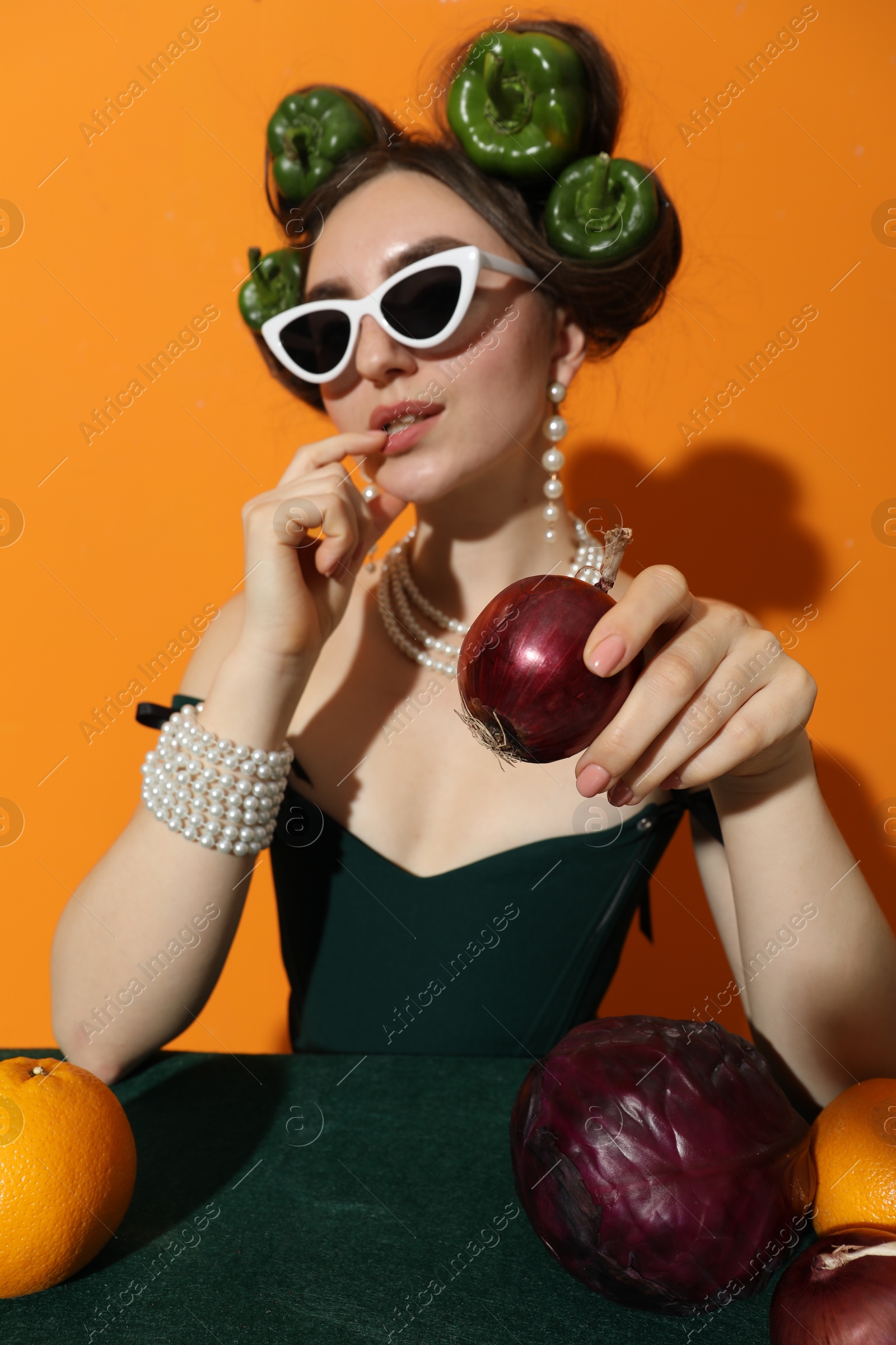 Photo of Young woman with peppers in her hair, sunglasses, fruits and vegetables at green table on orange background