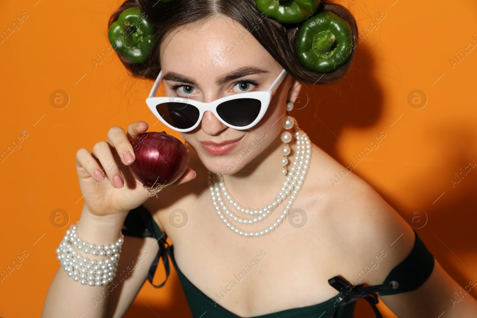 Photo of Young woman with peppers in her hair, sunglasses and onion on orange background