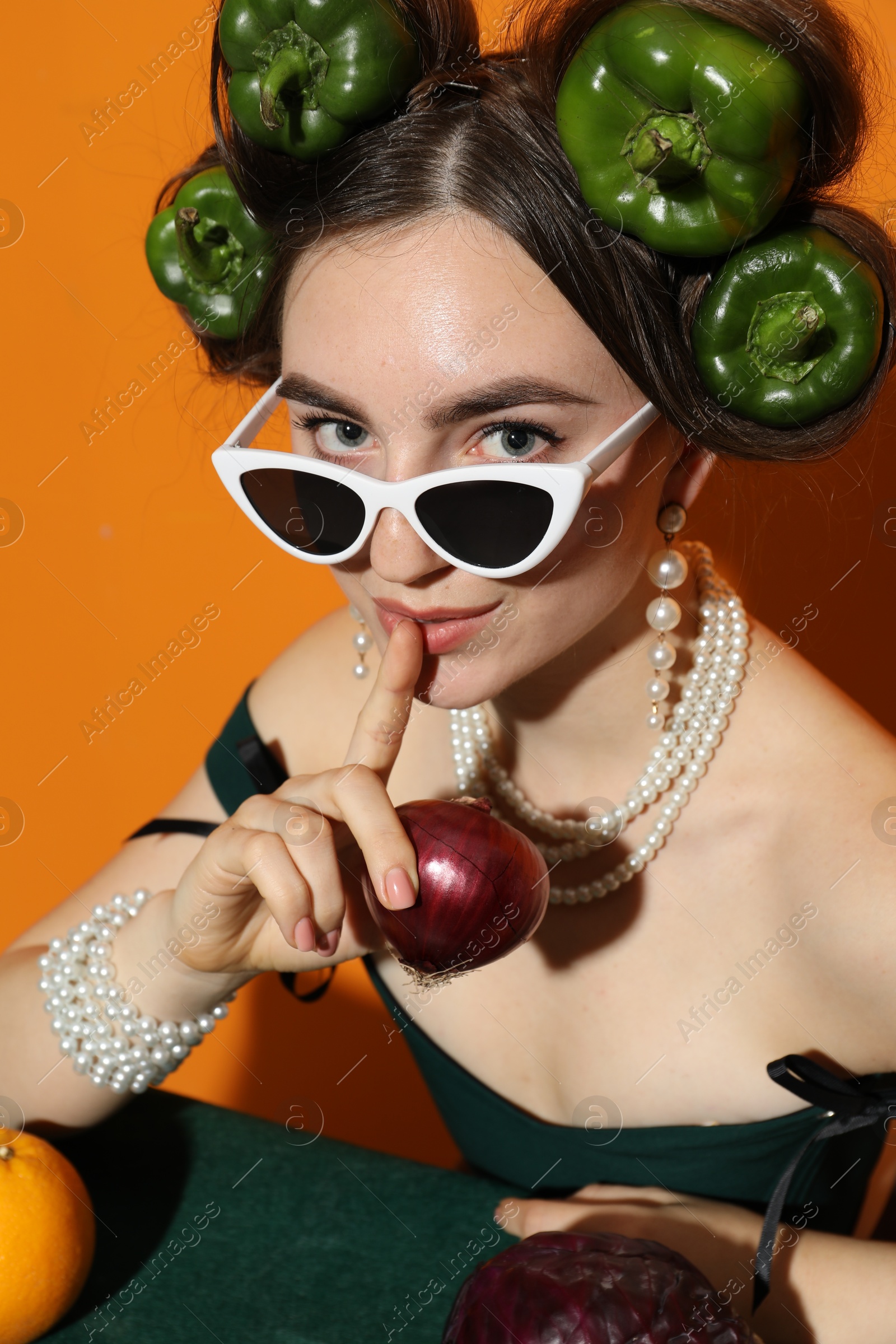 Photo of Young woman with peppers in her hair, sunglasses, fruits and vegetables at green table on orange background