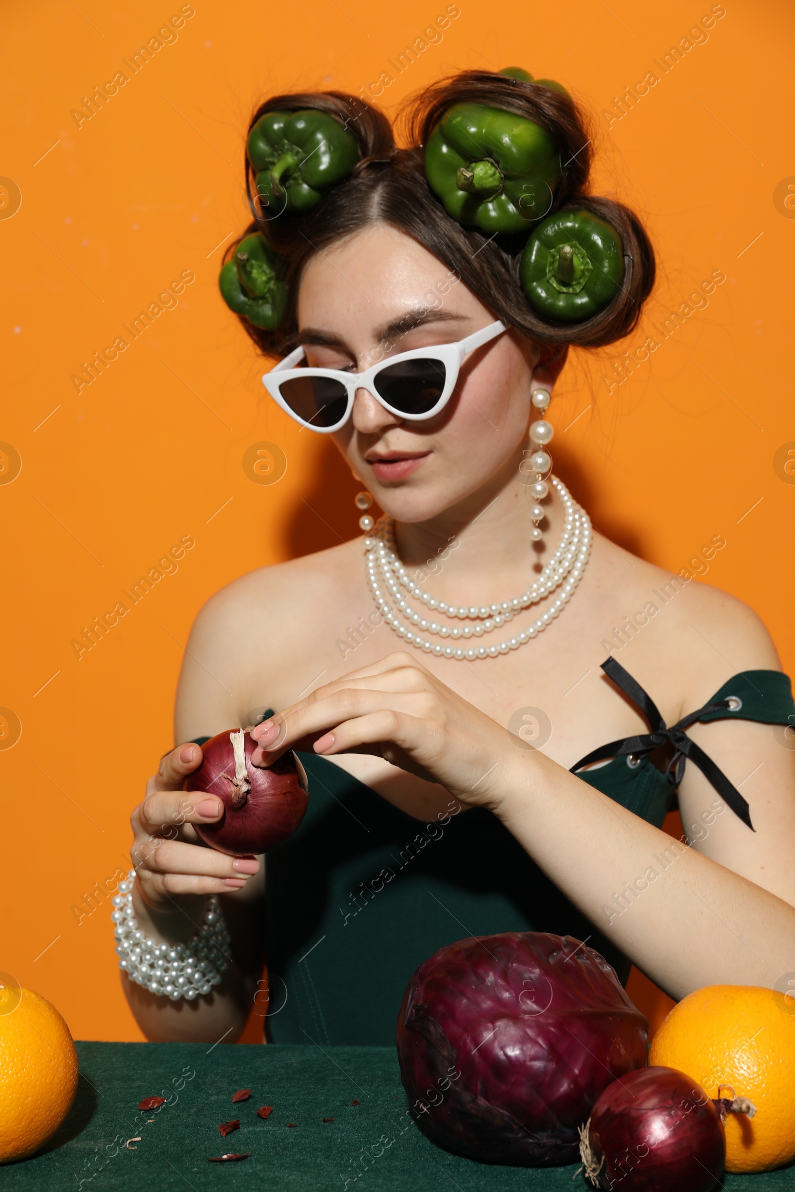 Photo of Young woman with peppers in her hair, sunglasses, fruits and vegetables at green table on orange background