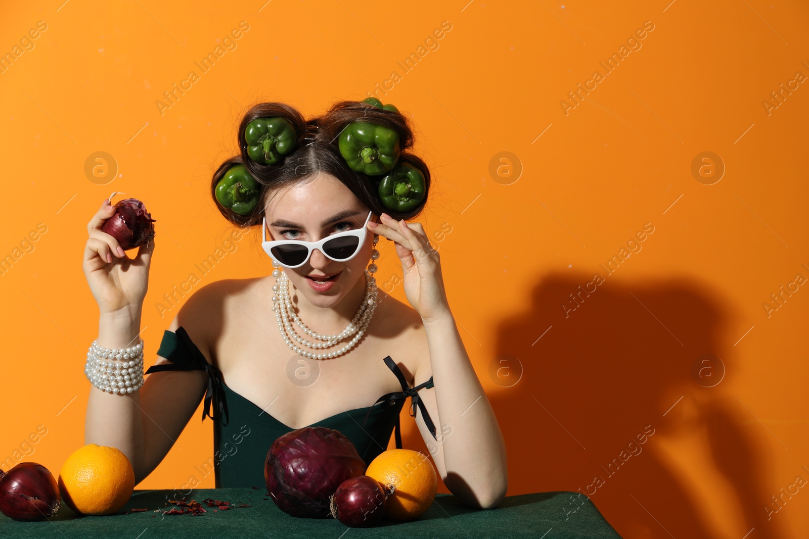 Photo of Young woman with peppers in her hair, sunglasses, fruits and vegetables at green table on orange background, space for text