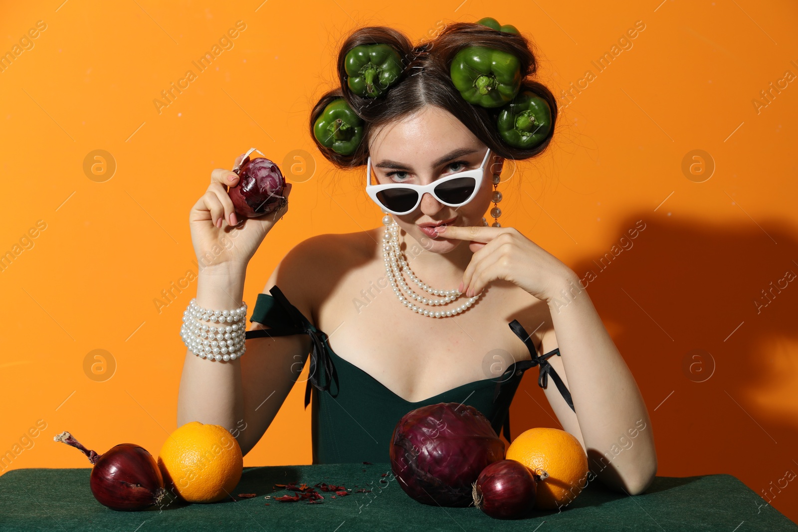 Photo of Young woman with peppers in her hair, sunglasses, fruits and vegetables at green table on orange background