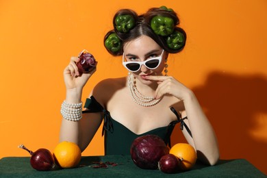 Photo of Young woman with peppers in her hair, sunglasses, fruits and vegetables at green table on orange background