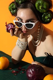 Photo of Young woman with peppers in her hair, sunglasses, fruits and vegetables at green table on orange background