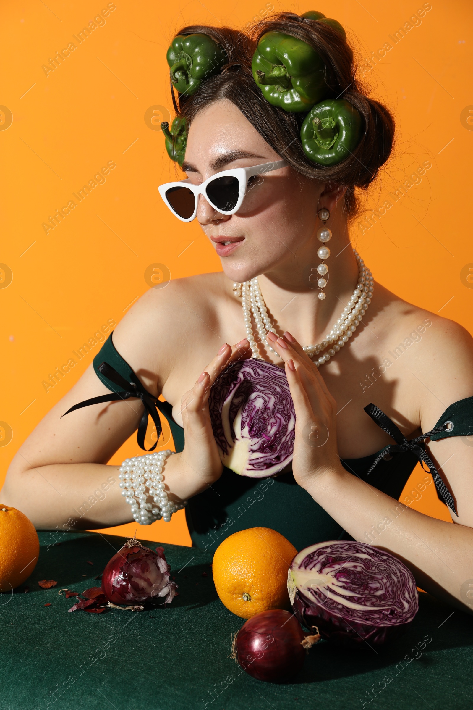 Photo of Young woman with peppers in her hair, sunglasses, fruits and vegetables at green table on orange background