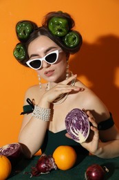 Young woman with peppers in her hair, sunglasses, fruits and vegetables at green table on orange background