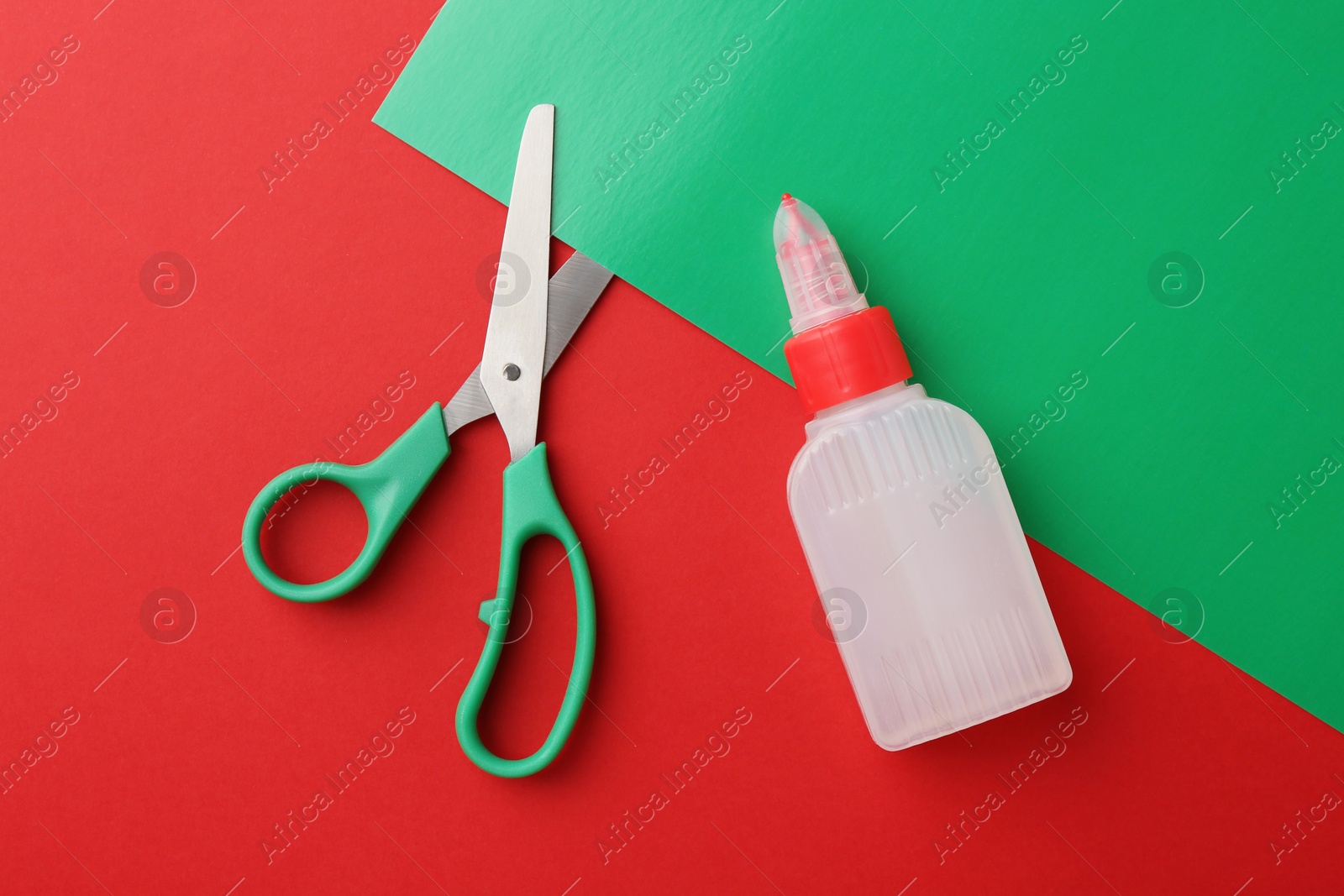 Photo of Bottle of glue, scissors and green paper on red background, flat lay