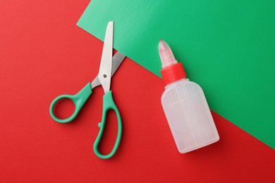 Photo of Bottle of glue, scissors and green paper on red background, flat lay