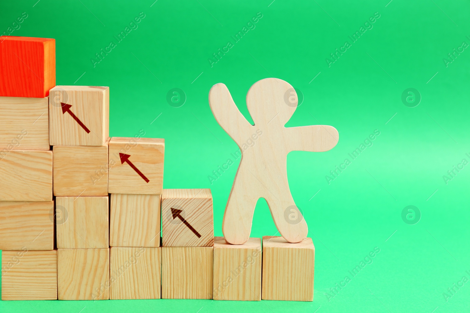 Image of Human figure on stair made of wooden blocks against green background. Cubes with arrows showing way to red one. Steps to success concept