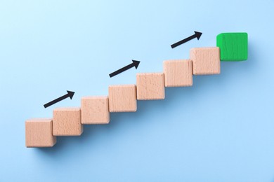 Image of Wooden cubes and last green one on light blue background, top view. Arrows showing direction to goal. Steps to success concept