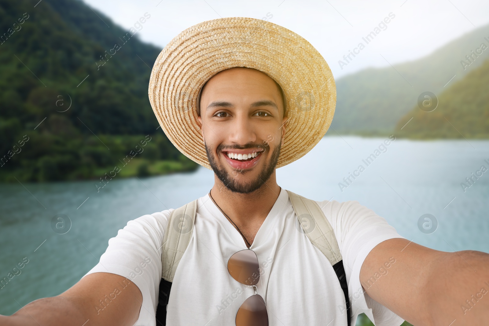 Image of Smiling young man in straw hat taking selfie near lake