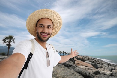 Smiling young man in straw hat taking selfie near sea