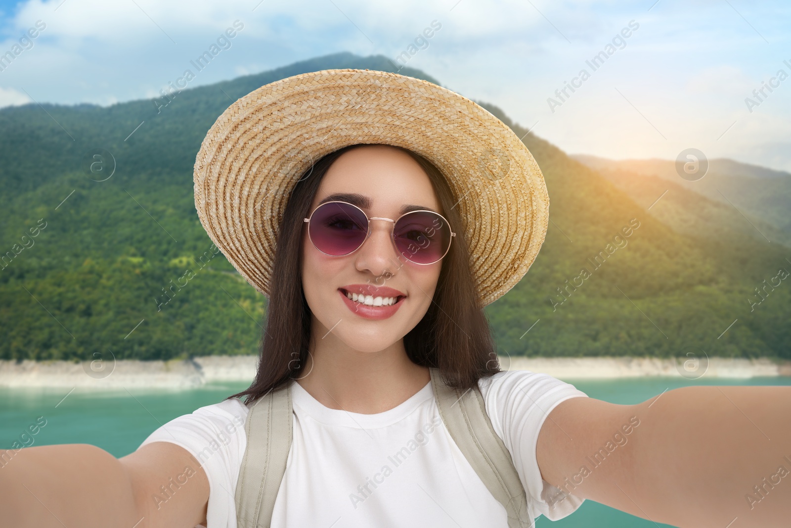 Image of Happy woman in straw hat taking selfie against lake and mountains