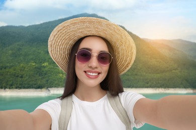 Happy woman in straw hat taking selfie against lake and mountains