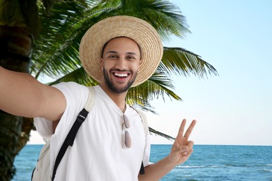 Image of Smiling young man in straw hat taking selfie near sea