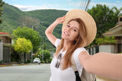 Image of Happy woman in straw hat taking selfie in mountain city