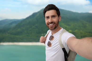 Image of Happy man with sunglasses taking selfie against lake and mountain