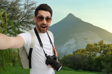 Image of Happy man with sunglasses and camera taking selfie in mountains