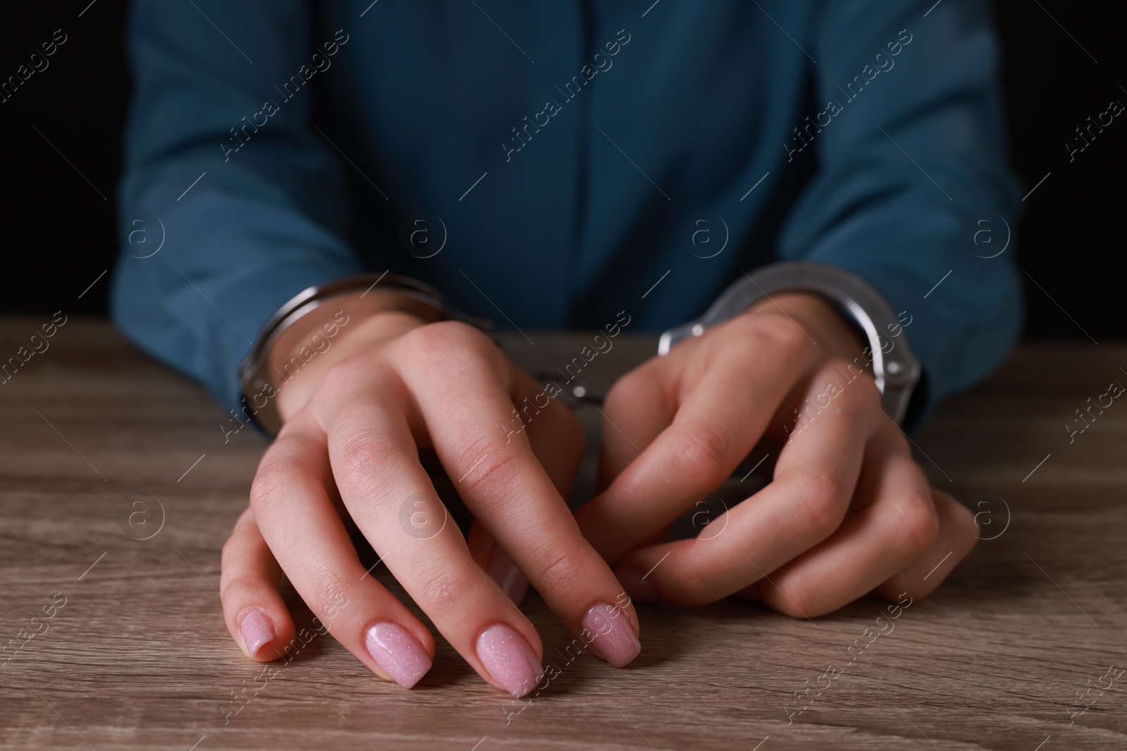 Photo of Woman in metal handcuffs at wooden table against black background, closeup