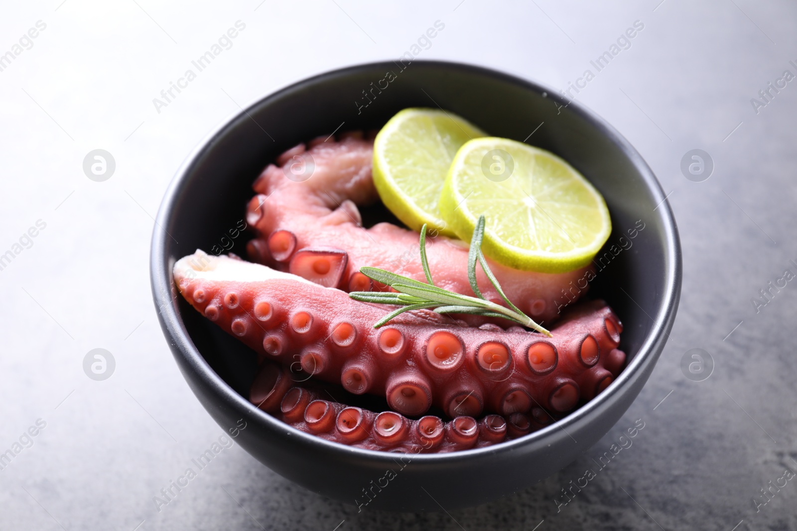 Photo of Fresh raw octopus, lemon and rosemary in bowl on grey table, closeup