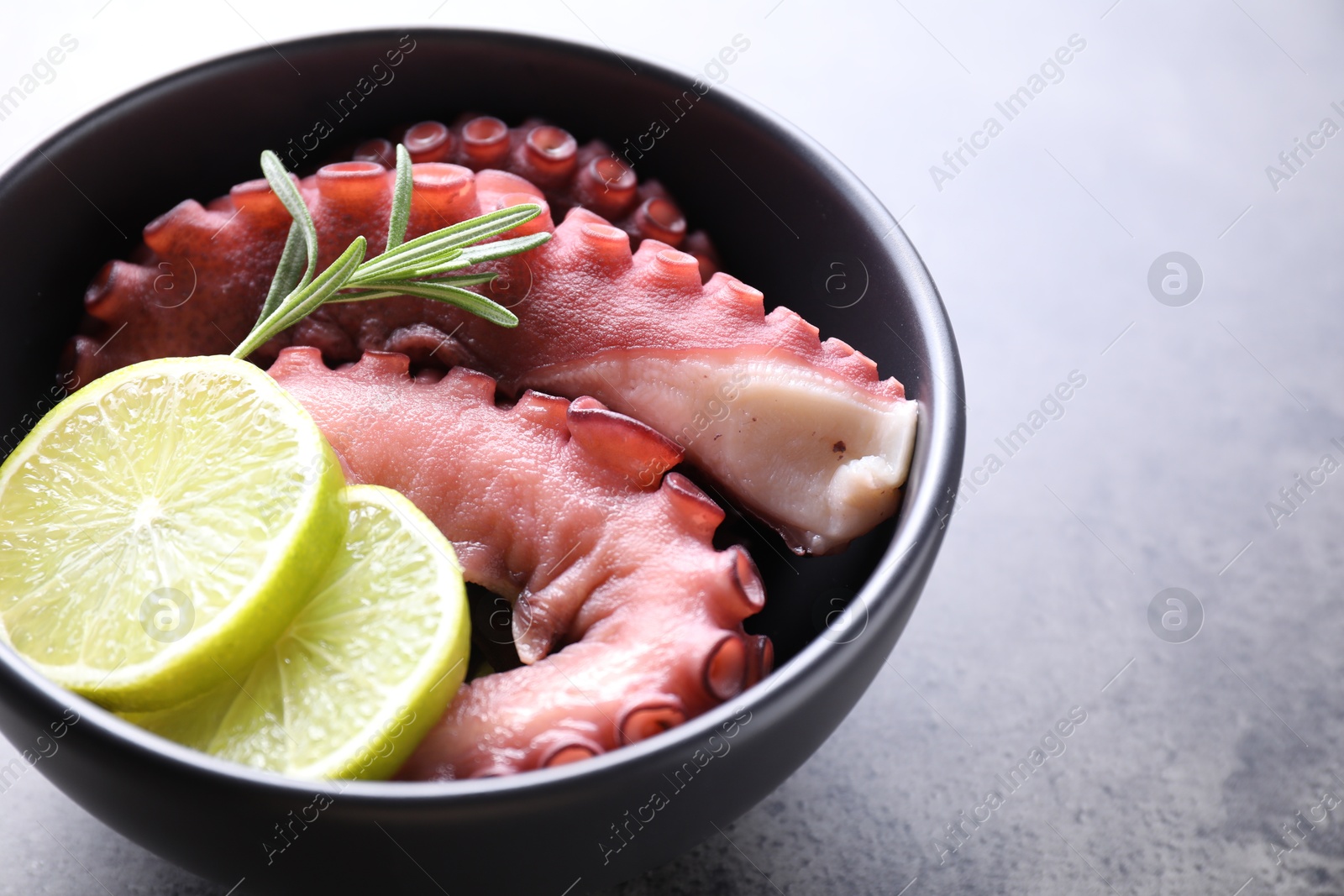 Photo of Fresh raw octopus, lemon and rosemary in bowl on grey table, closeup