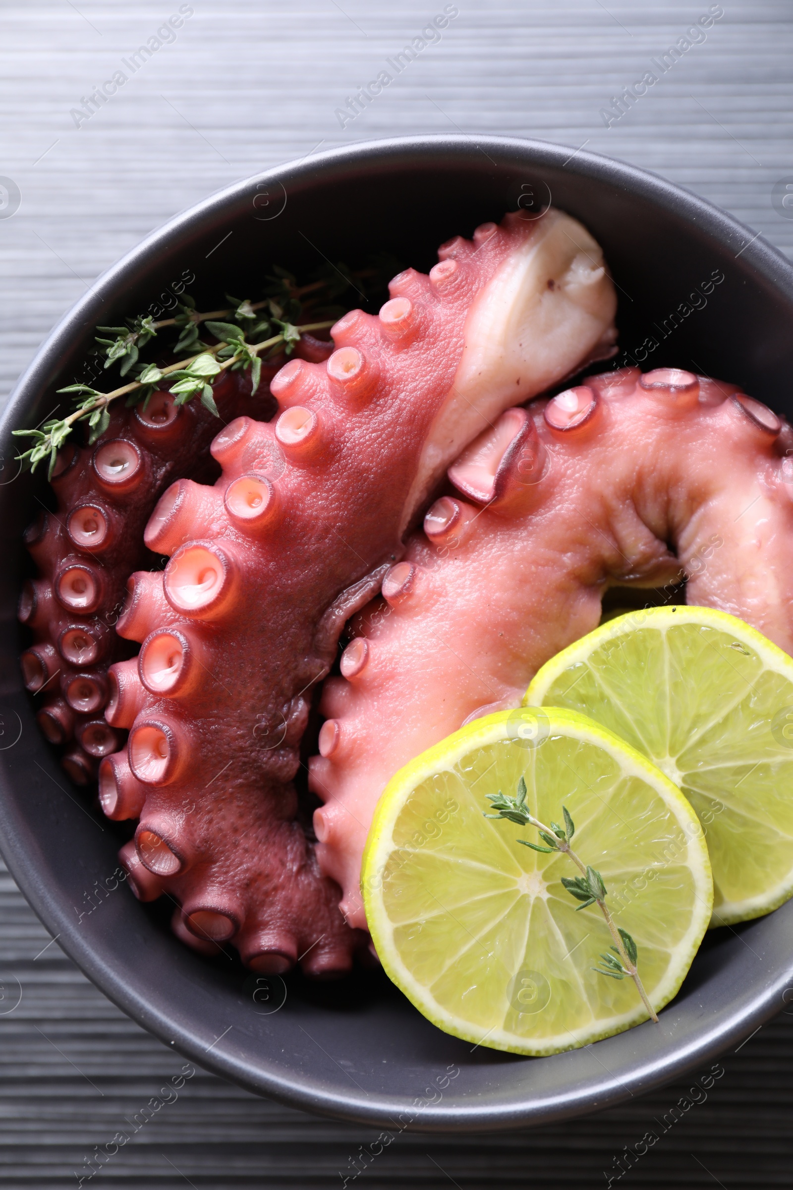 Photo of Fresh raw octopus, lemon and thyme in bowl on dark textured table, top view