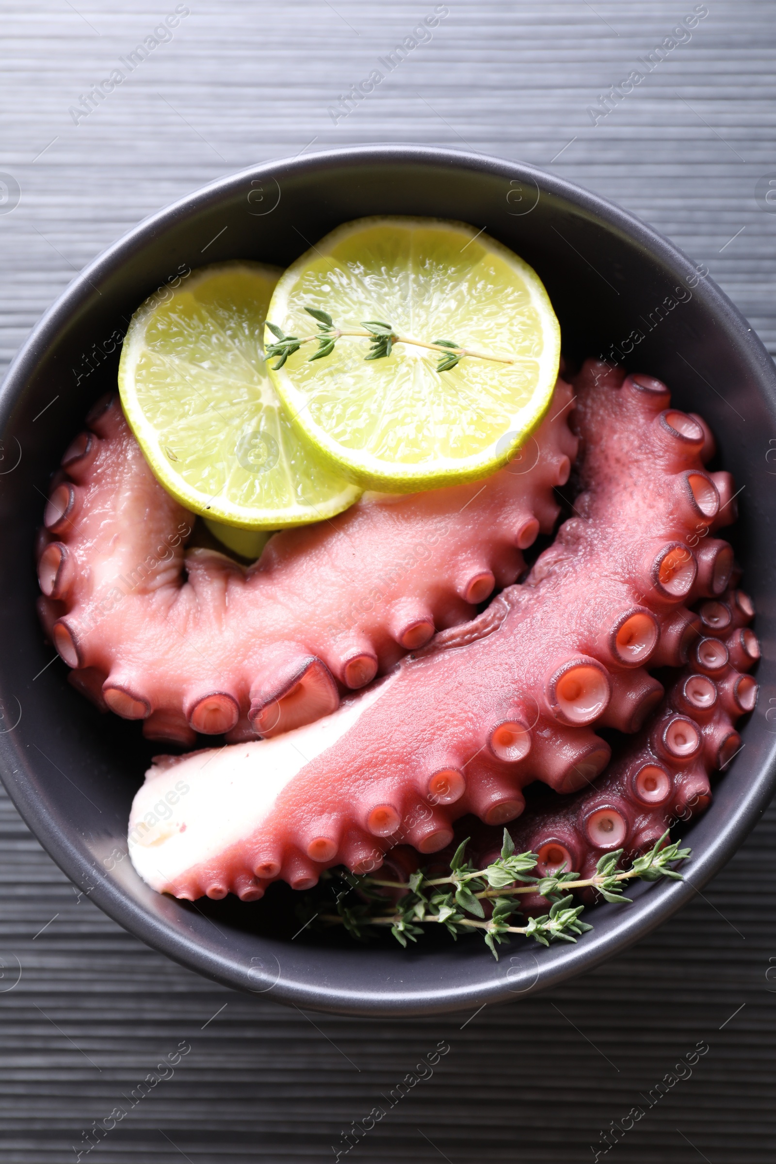 Photo of Fresh raw octopus, lemon and thyme in bowl on dark textured table, top view