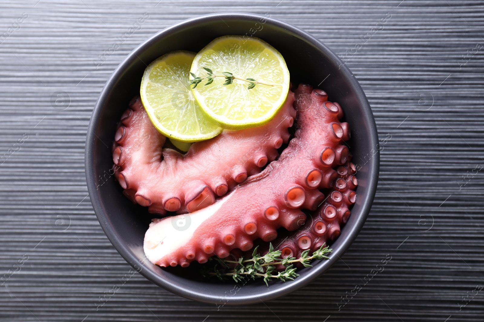 Photo of Fresh raw octopus, lemon and thyme in bowl on dark textured table, top view