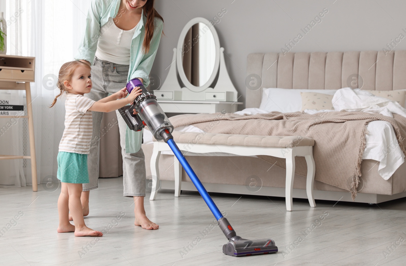 Photo of Young woman and her daughter cleaning floor with cordless vacuum cleaner in bedroom, closeup