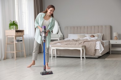 Photo of Smiling young woman cleaning floor with cordless vacuum cleaner while her daughter sleeping in bedroom