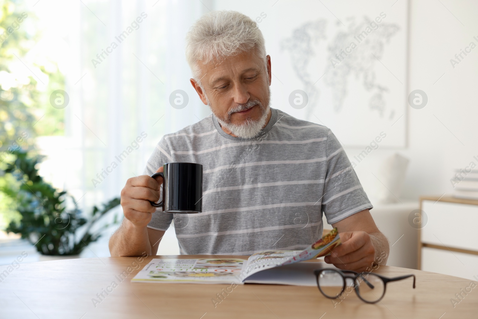 Photo of Senior man with cup of drink reading magazine at table indoors