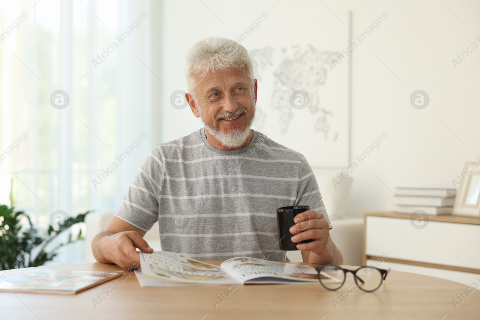 Photo of Senior man with cup of drink reading magazine at table indoors