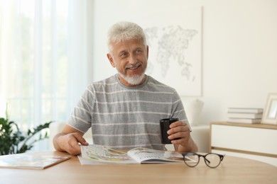 Senior man with cup of drink reading magazine at table indoors