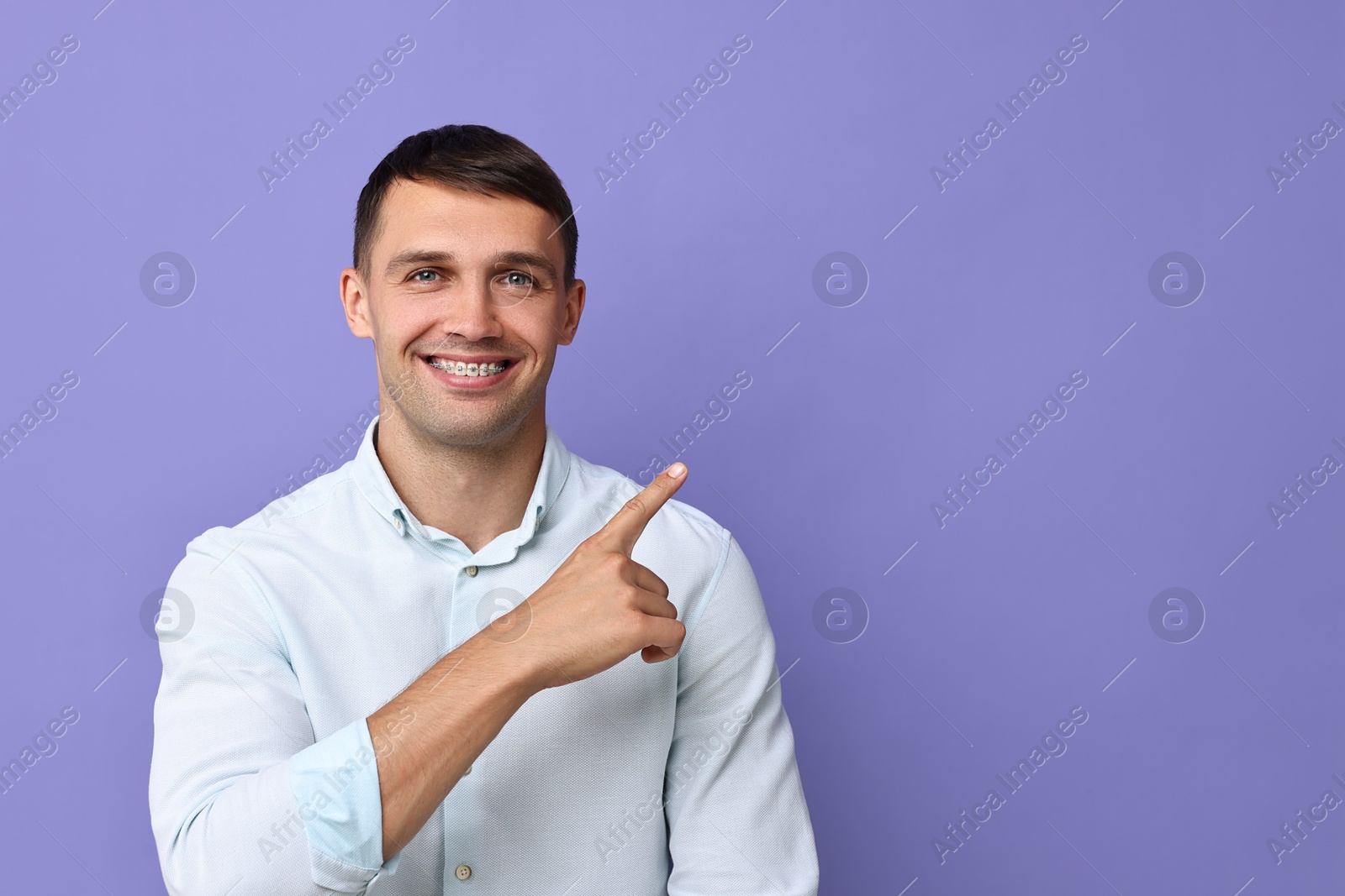 Photo of Smiling man with dental braces pointing at something on violet background. Space for text