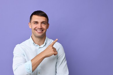 Photo of Smiling man with dental braces pointing at something on violet background. Space for text