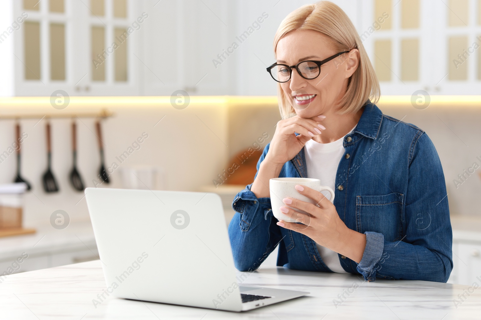 Photo of Happy middle aged woman with cup of drink using laptop at white marble table in kitchen