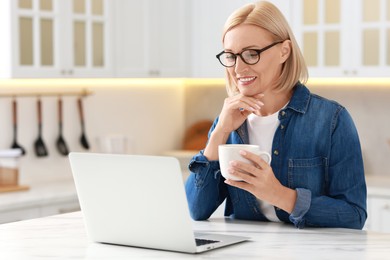 Happy middle aged woman with cup of drink using laptop at white marble table in kitchen