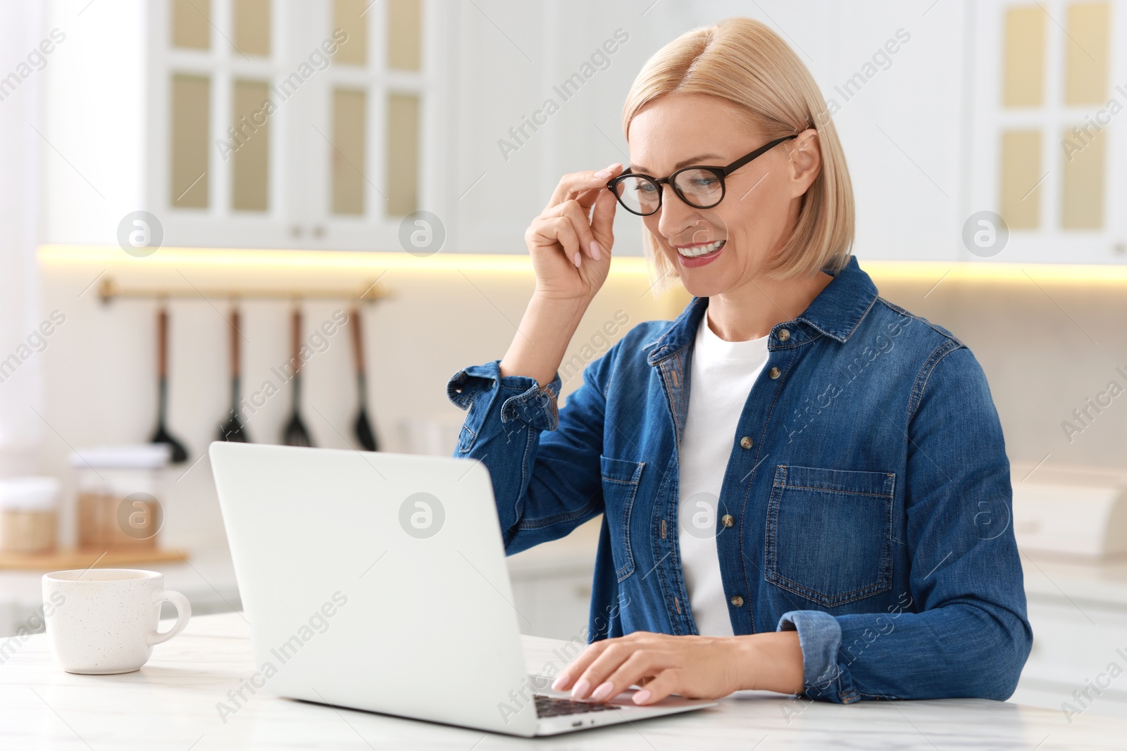 Photo of Happy middle aged woman using laptop at white marble table in kitchen