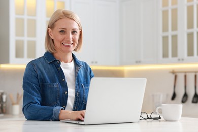 Happy middle aged woman using laptop at white marble table in kitchen