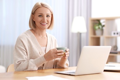 Happy middle aged woman with cup of drink working at table indoors