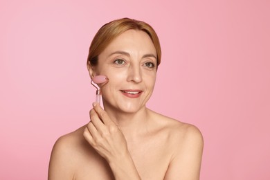 Photo of Smiling woman doing facial self massage with roller on pink background