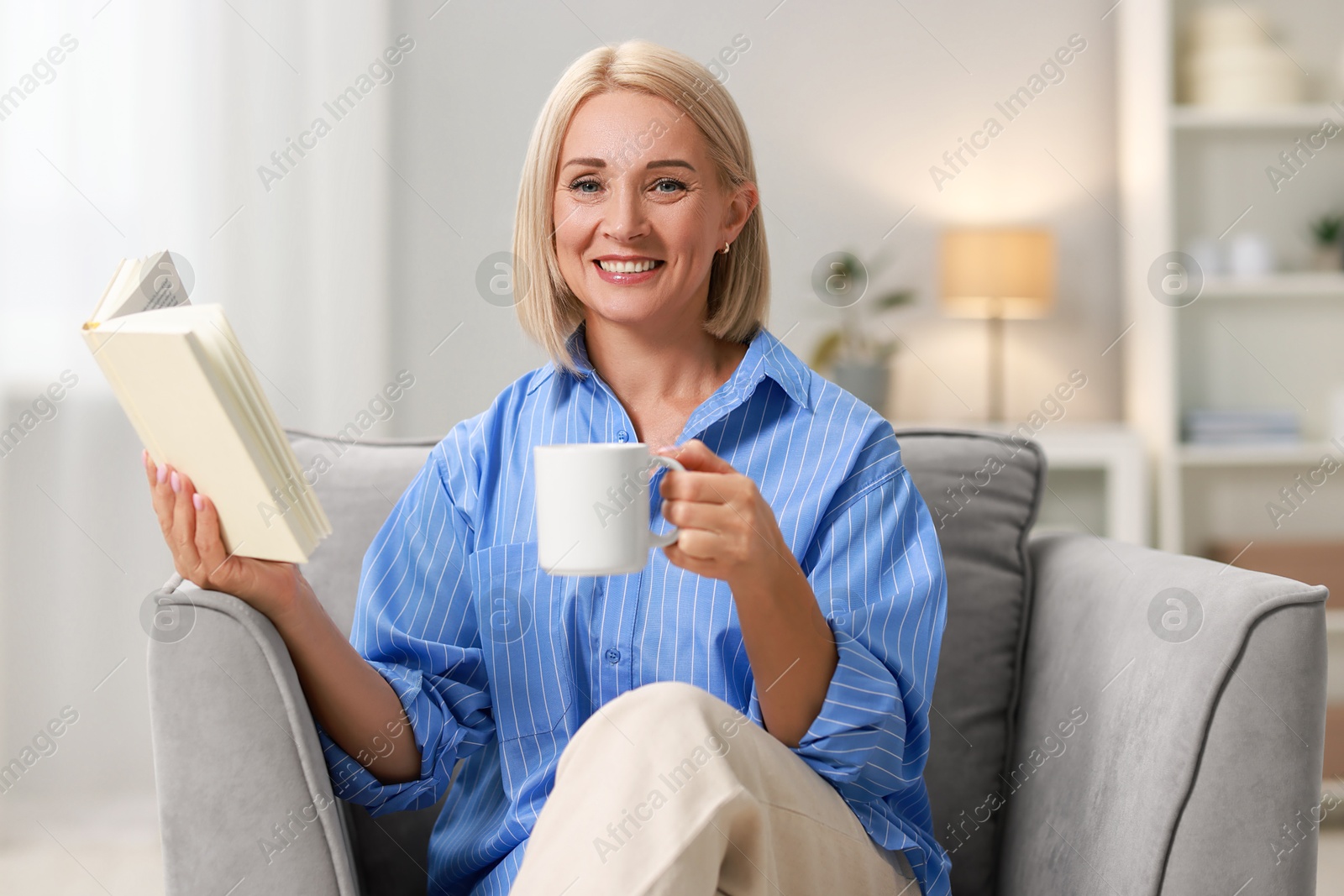 Photo of Smiling middle aged woman with cup of hot drink reading book at home