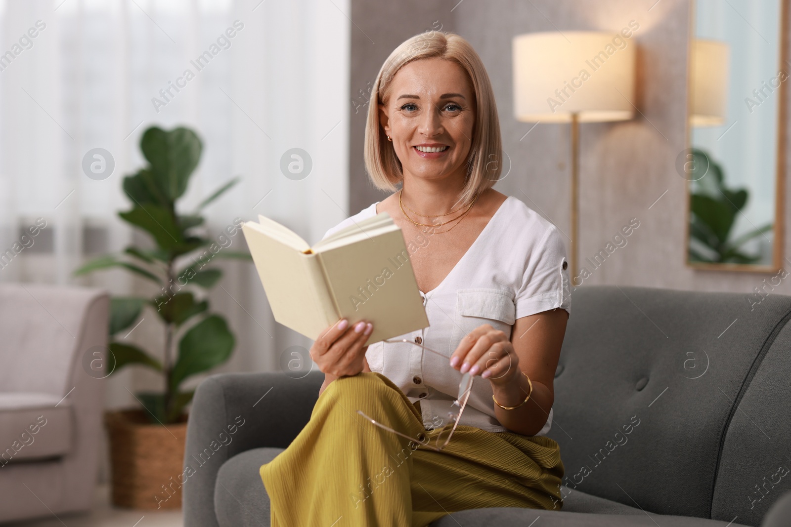 Photo of Portrait of smiling middle aged woman with glasses reading book on sofa at home