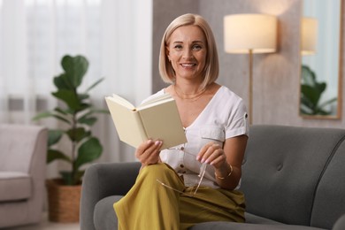 Photo of Portrait of smiling middle aged woman with glasses reading book on sofa at home