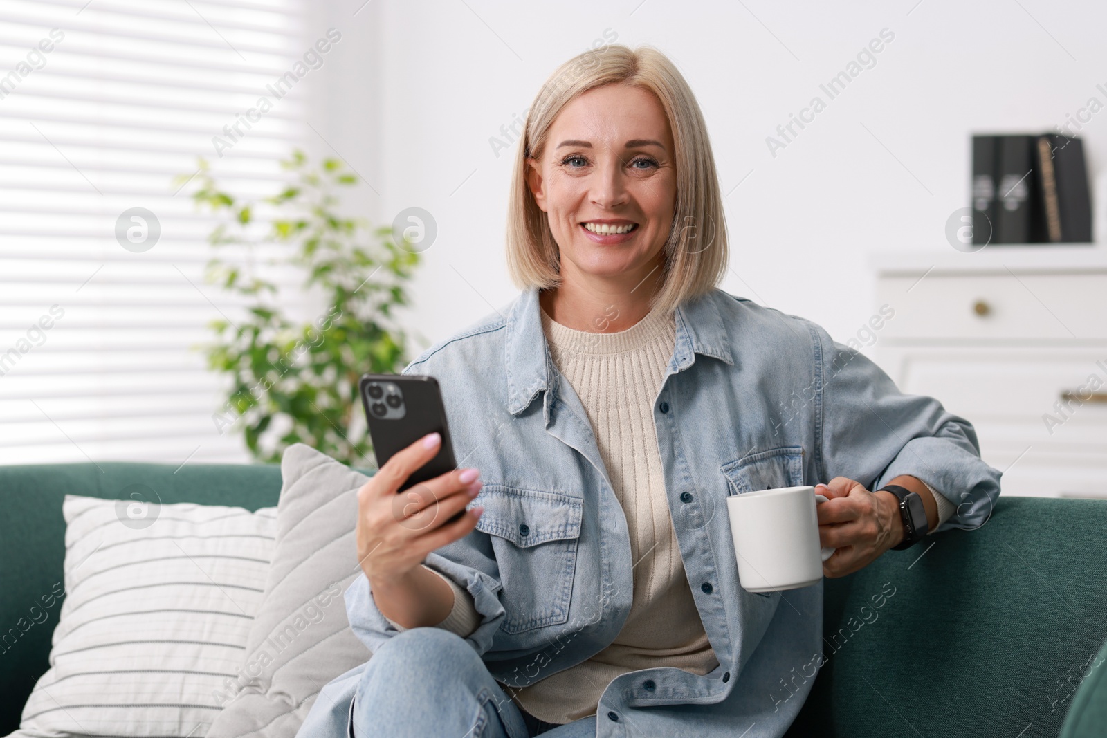 Photo of Smiling middle aged woman with cup of hot drink using smartphone at home