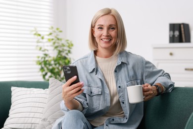 Smiling middle aged woman with cup of hot drink using smartphone at home