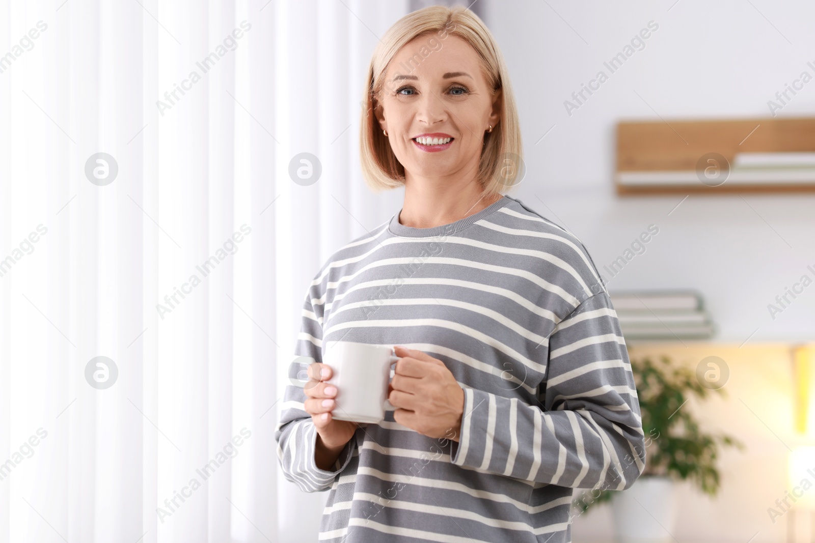 Photo of Smiling middle aged woman with cup of hot drink near window at home