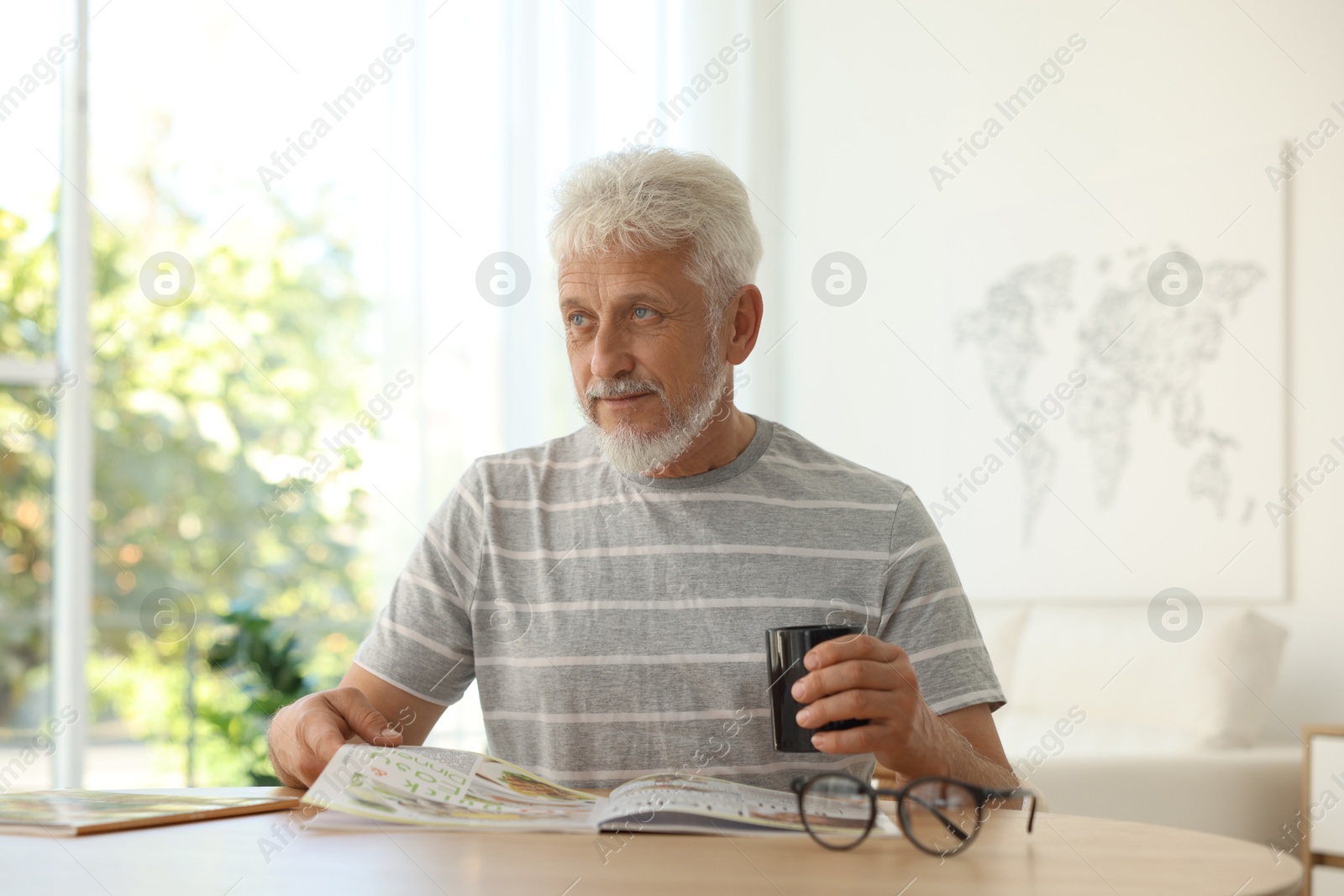 Photo of Senior man with cup of drink reading magazine at table indoors