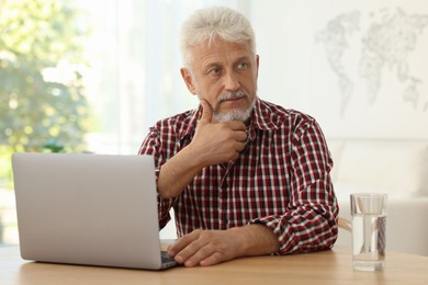 Senior man with laptop and glass of water at table indoors